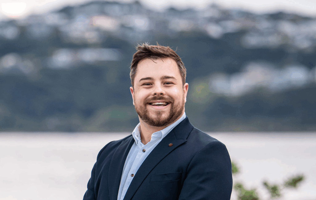 Josh Trlin profile photo Josh Trlin smiling in a suit jacket with a blurred coastal background.