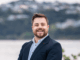 Josh Trlin smiling in a suit jacket with a blurred coastal background.