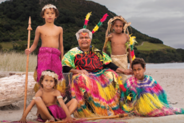 Celebrating Bislama Language and Culture A Ni-Vanuatu elder in traditional dress sits on the beach with five children in colourful cultural attire, against a backdrop of coastal hills.