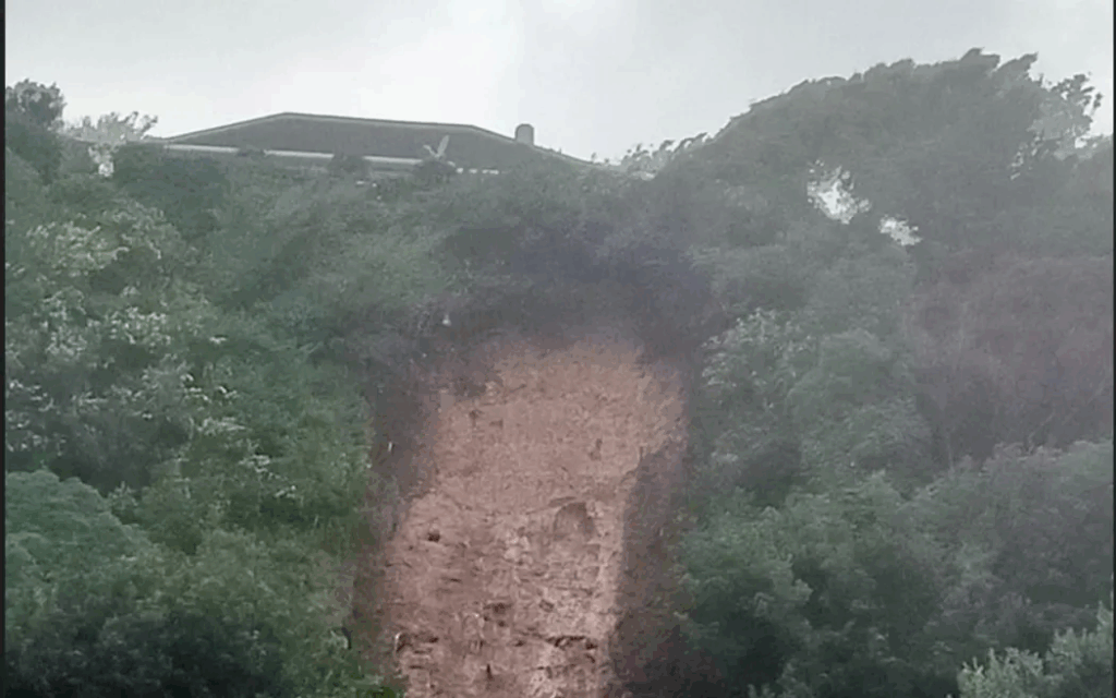 Landslip After Heavy Rain Hits Porirua Homes Muddy landslip debris piled against the rear walls of suburban houses in Porirua, with emergency cones and tape visible nearby.