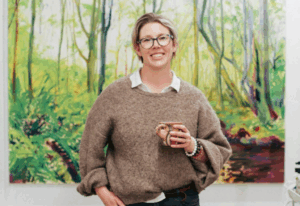 Alicja Gear, art tutor, standing in front of a colourful painting holding a cup.