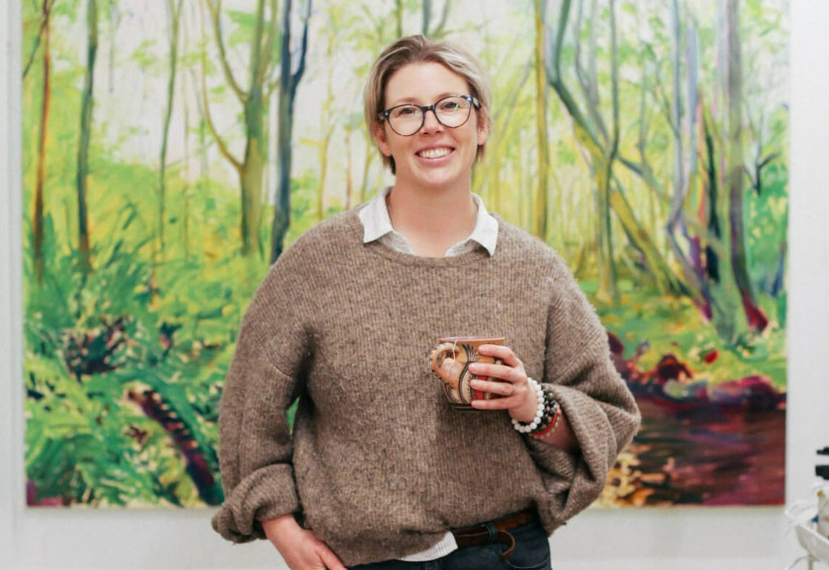 Alicja Gear at Mana Arts Society Alicja Gear, art tutor, standing in front of a colourful painting holding a cup.