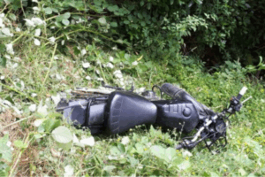 A black dirt bike lying on its side in dense green bush and undergrowth, partially obscured by plants, indicating an off-track crash or abandonment.