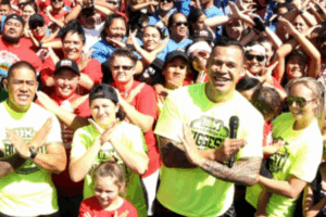 Dave Letele leads a large crowd wearing BBM shirts and making an X with their arms during a community event.