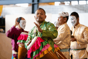 A group of men in traditional Southeast Asian attire perform with colourful drums and masks during a cultural celebration.