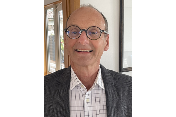 Grenville Gaskell wearing glasses and a checked shirt with a grey blazer, smiling indoors near a window.