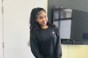 Teenage girl in black Nike sweatshirt, smiling and standing indoors.
