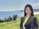 Yan Zang standing in front of a coastal landscape on a sunny day, smiling and wearing a dark green jacket with a cream blouse.
