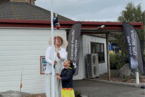 An adult in traditional Cook Islands attire and a child raise the Cook Islands flag outside a building with Central Pacific Collective banners.