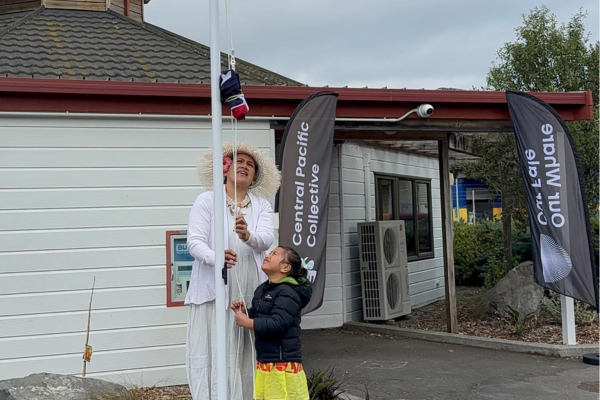 An adult in traditional Cook Islands attire and a child raise the Cook Islands flag outside a building with Central Pacific Collective banners.