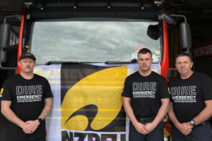 Three firefighters stand in front of a fire engine wearing "Dire Emergency" union shirts, protesting for better resourcing and support from Fire and Emergency NZ.