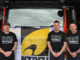 Three firefighters stand in front of a fire engine wearing "Dire Emergency" union shirts, protesting for better resourcing and support from Fire and Emergency NZ.