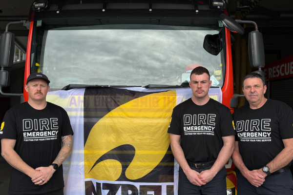 Three firefighters stand in front of a fire engine wearing "Dire Emergency" union shirts, protesting for better resourcing and support from Fire and Emergency NZ.