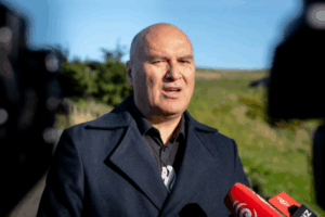 Helmut Modlik, Chief Executive of Ngāti Toa Rangatira, speaks outdoors at a media event. He is wearing a dark coat and standing in front of grassy hills, with red RNZ microphones visible in the foreground.