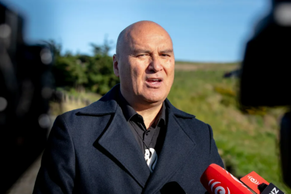 Helmut Modlik – Chief Executive of Ngāti Toa Rangatira Helmut Modlik, Chief Executive of Ngāti Toa Rangatira, speaks outdoors at a media event. He is wearing a dark coat and standing in front of grassy hills, with red RNZ microphones visible in the foreground.
