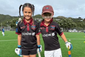 Two children wearing North City Cricket Club uniforms smile on a cricket field with trees and hills in the background