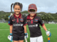 Two children wearing North City Cricket Club uniforms smile on a cricket field with trees and hills in the background