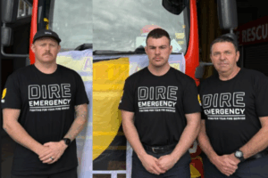 Three firefighters wearing “Dire Emergency” union shirts stand in front of a fire truck draped with an NZPFU banner, preparing for a strike to raise awareness about working conditions.