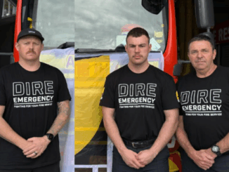 Three firefighters wearing “Dire Emergency” union shirts stand in front of a fire truck draped with an NZPFU banner, preparing for a strike to raise awareness about working conditions.