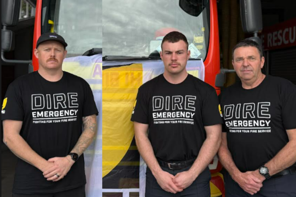 Three firefighters wearing “Dire Emergency” union shirts stand in front of a fire truck draped with an NZPFU banner, preparing for a strike to raise awareness about working conditions.