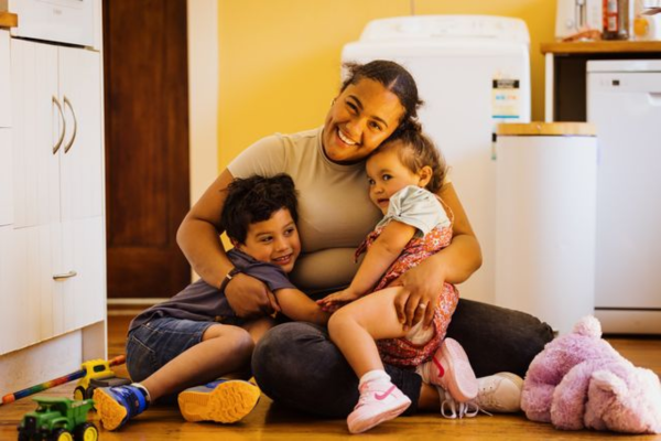 Mother hugs her two young children on the floor of a warm family home in Porirua.