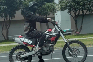 A person wearing a black helmet and face covering rides a black and red dirt bike with the number 450 on the side along a city street in Porirua.