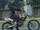 A person wearing a black helmet and face covering rides a black and red dirt bike with the number 450 on the side along a city street in Porirua.