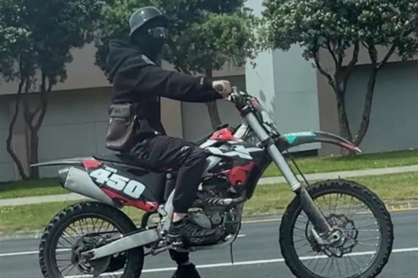 A person wearing a black helmet and face covering rides a black and red dirt bike with the number 450 on the side along a city street in Porirua.