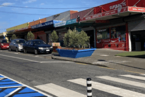 A row of small Porirua shops with colourful signage, parked cars, a pedestrian crossing, and a large blue planter on the footpath.