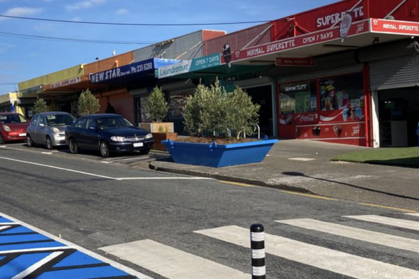 A row of small Porirua shops with colourful signage, parked cars, a pedestrian crossing, and a large blue planter on the footpath.