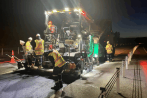 Night-time roadworks underway on Transmission Gully, with workers operating paving machinery under floodlights while traffic lanes are closed.