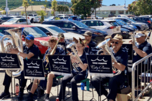 Members of Porirua City Brass and Porirua Youth Band perform together outside The Warehouse, seated with brass instruments as shoppers and parked cars fill the background.