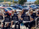 Members of Porirua City Brass and Porirua Youth Band perform together outside The Warehouse, seated with brass instruments as shoppers and parked cars fill the background.