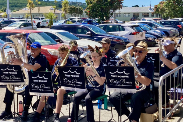 Members of Porirua City Brass and Porirua Youth Band perform together outside The Warehouse, seated with brass instruments as shoppers and parked cars fill the background.