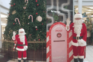 Santa Claus stands beside a North Pole-themed post box display at North City Shopping Centre, waving in front of a large decorated Christmas tree.