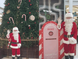 Santa Claus stands beside a North Pole-themed post box display at North City Shopping Centre, waving in front of a large decorated Christmas tree.