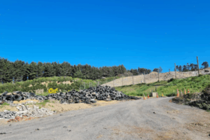 Gravel access road leading through a partially filled landfill site with earth embankments and vegetation under a clear blue sky.