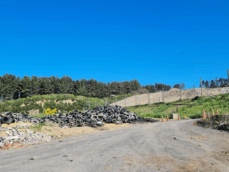 Gravel access road leading through a partially filled landfill site with earth embankments and vegetation under a clear blue sky.