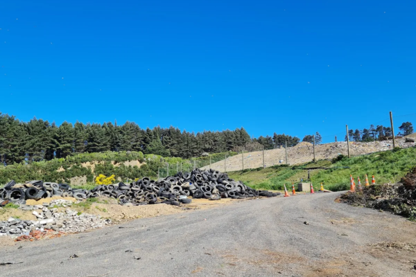 Gravel access road leading through a partially filled landfill site with earth embankments and vegetation under a clear blue sky.