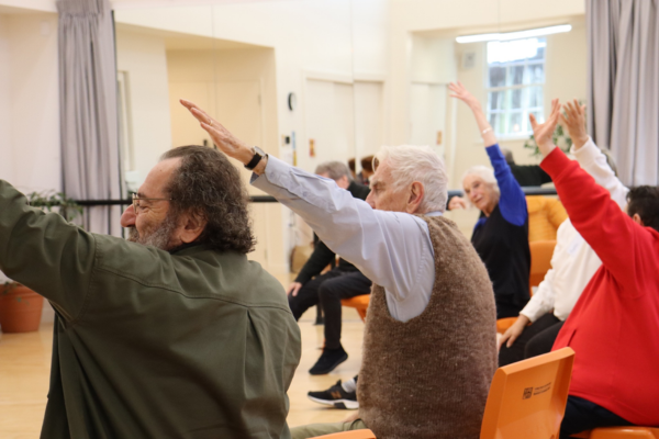 A group of older adults seated in chairs raise one arm during a gentle exercise class in a community hall.