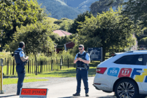 Two Police officers stand beside a marked patrol car at a road cordon on Paekākāriki Hill, with rural fencing, trees and bush-covered hills in the background.