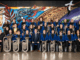 Members of Porirua City Brass pose for a formal group photo, wearing blue jackets and holding brass instruments.