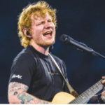 A male musician sings into a microphone while playing an acoustic guitar on a brightly lit stage.