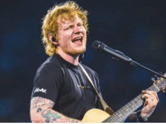 A male musician sings into a microphone while playing an acoustic guitar on a brightly lit stage.