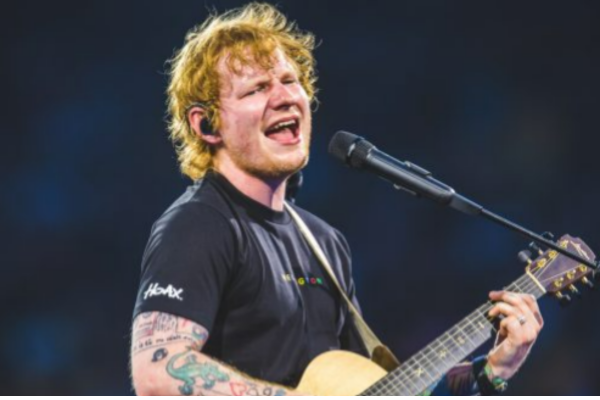 A male musician sings into a microphone while playing an acoustic guitar on a brightly lit stage.
