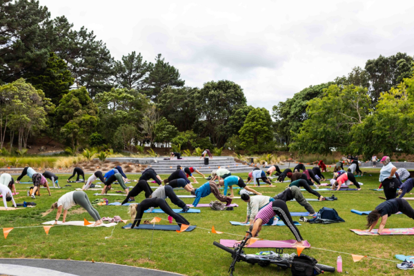People take part in an outdoor BodyBalance class on the grass at Aotea Lagoon, holding yoga-style poses on mats.