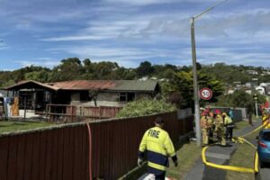 Firefighters stand on a Porirua street as hoses run toward a badly fire-damaged house, with the roof and windows visibly burned.