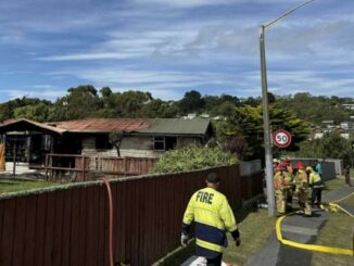 Firefighters stand on a Porirua street as hoses run toward a badly fire-damaged house, with the roof and windows visibly burned.
