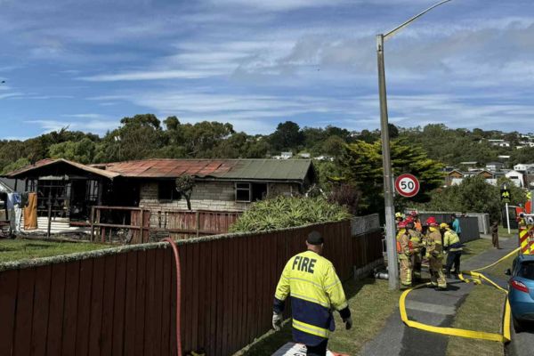 Firefighters stand on a Porirua street as hoses run toward a badly fire-damaged house, with the roof and windows visibly burned.