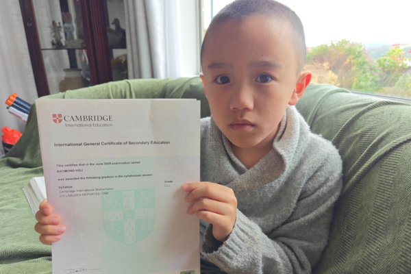 A young boy sits on a couch holding a Cambridge International Education certificate for mathematics.